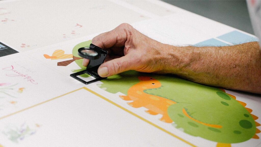 A person uses a magnifying tool to inspect a printed illustration of an orange dinosaur on a sheet with various colorful designs.