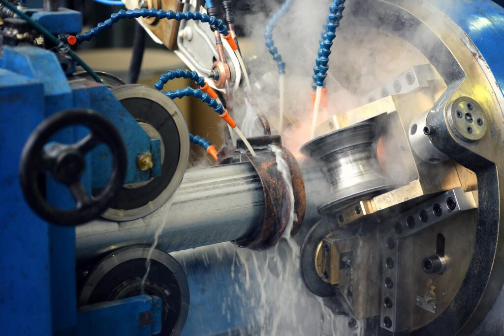A close-up of an industrial metalworking machine shaping a cylindrical metal piece, with coolant fluid being sprayed onto the work area, creating mist and steam. Blue and orange hoses direct the fluid onto the metal surface.
