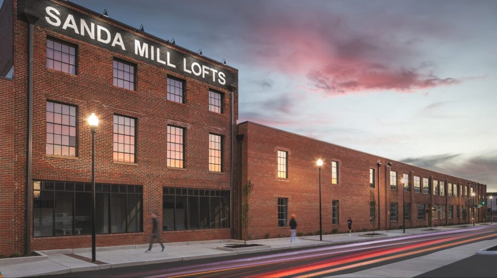 A large brick building with a sign reading "SANDA MILL LOFTS" is seen at dusk. Streetlights glow along the sidewalk, and a few blurred pedestrians walk by under a colorful evening sky.