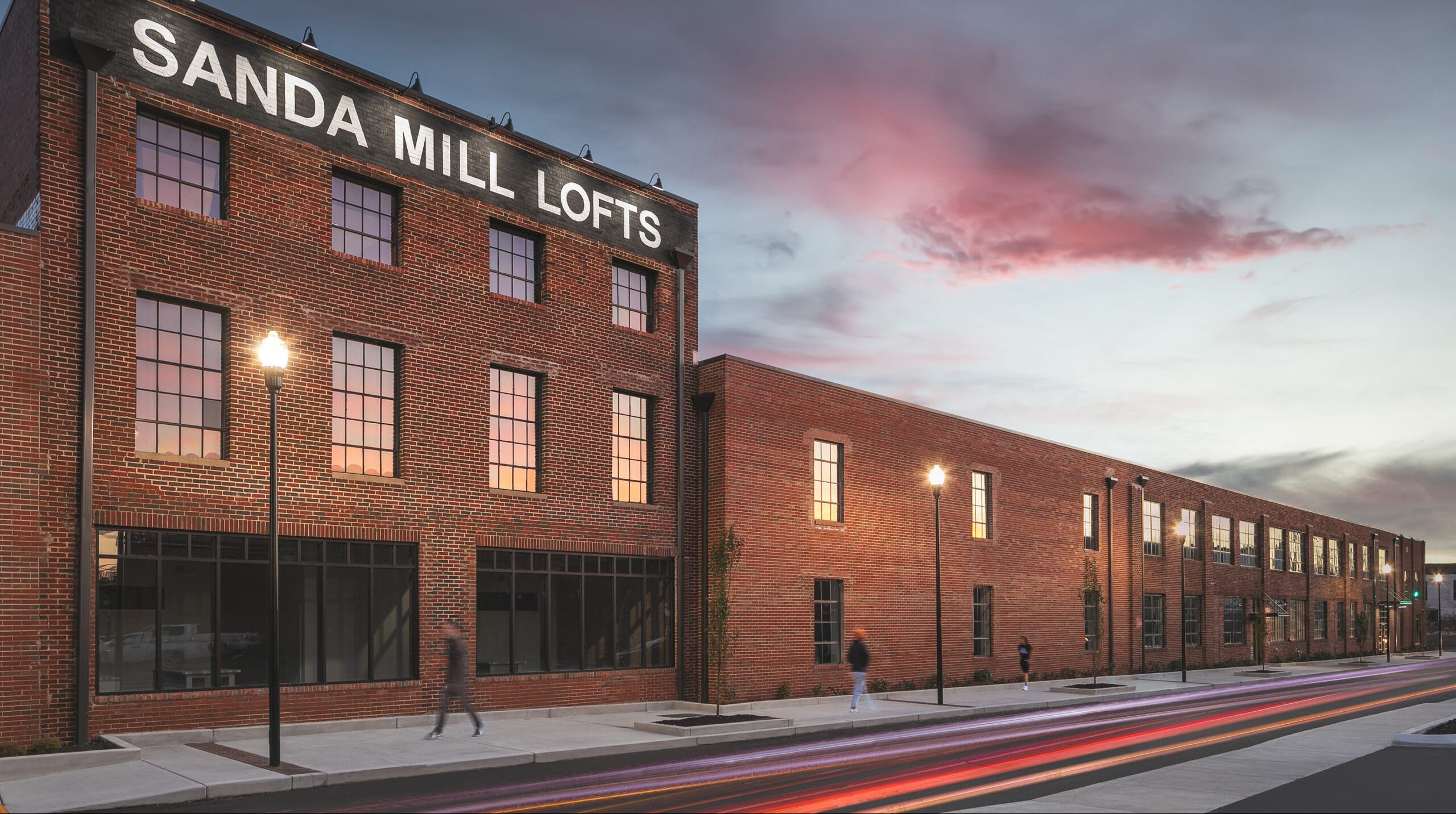 A large brick building with a sign reading "SANDA MILL LOFTS" is seen at dusk. Streetlights glow along the sidewalk, and a few blurred pedestrians walk by under a colorful evening sky.