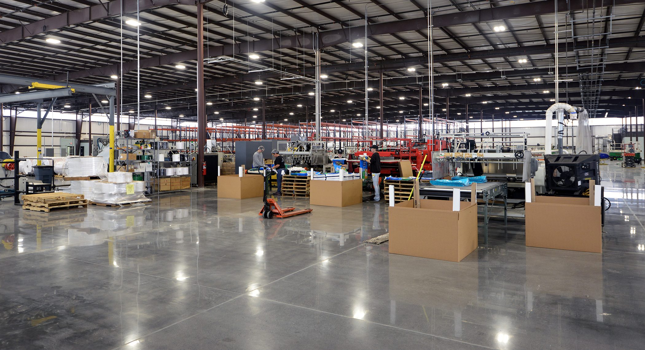 A spacious factory floor with machinery, cardboard boxes, wooden pallets, and workers operating equipment. The area is well-lit with high ceilings and polished concrete floors, creating an organized and clean industrial environment.