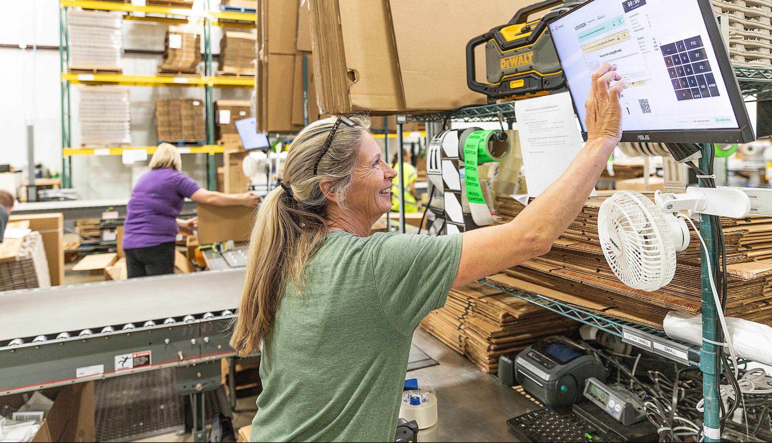A woman in a green shirt smiles while touching a screen at a workstation in a warehouse. Shelves with boxes and packing materials are in the background, and another person is working nearby.