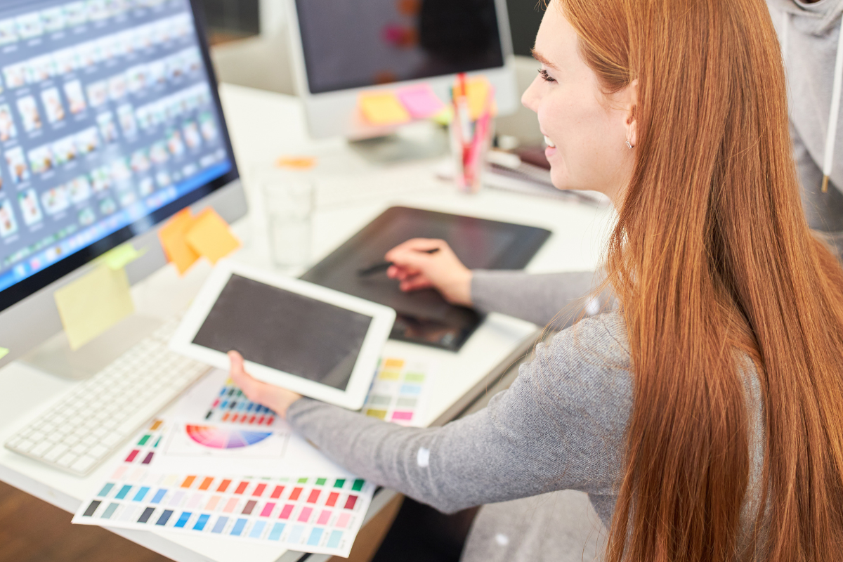 A woman with long red hair sits at a desk using a tablet and stylus, with a color swatch book and computer monitor displaying image thumbnails in front of her, suggesting she is working on a design project.