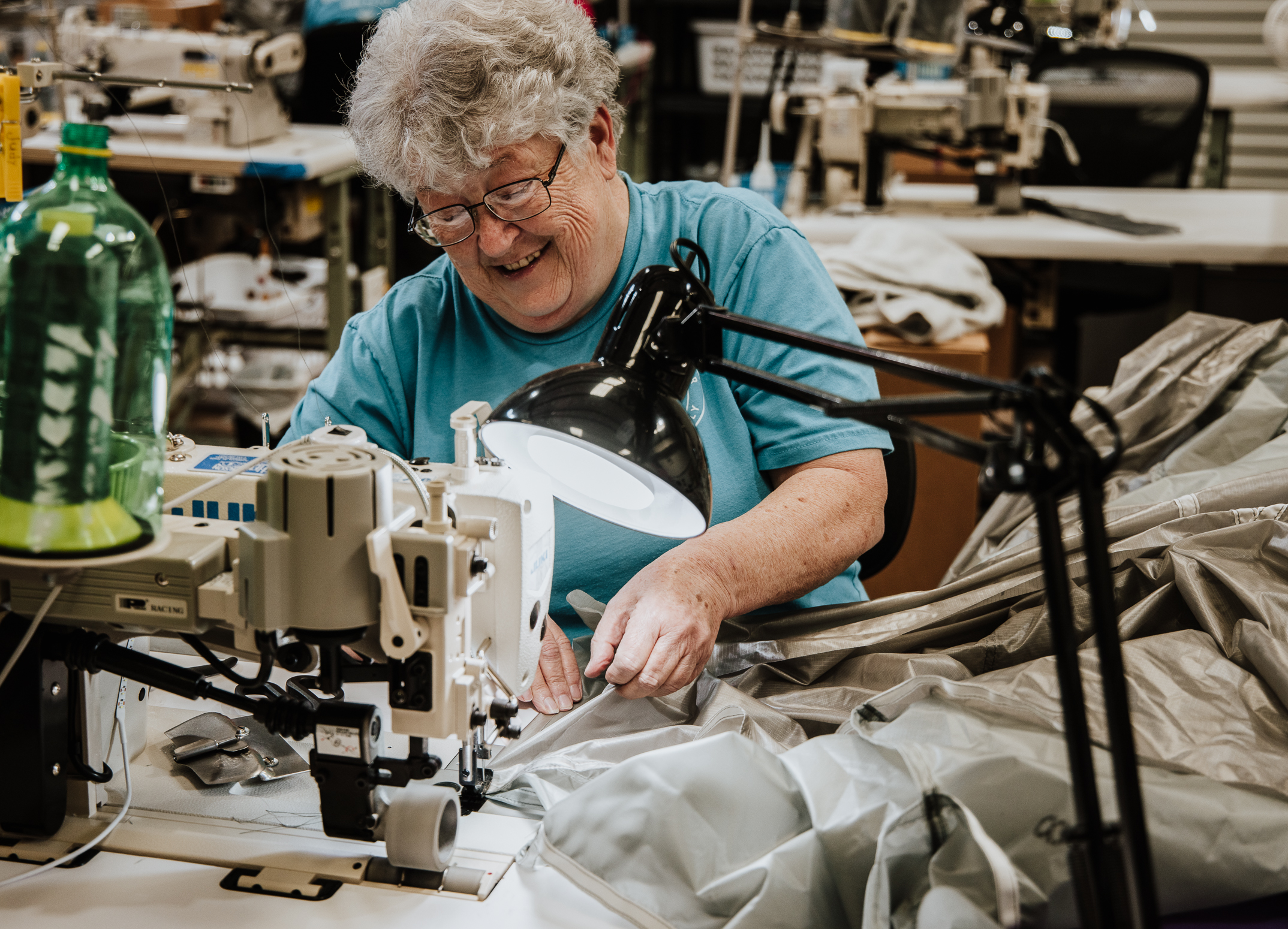 An older woman with curly gray hair smiles while sewing gray fabric with an industrial sewing machine in a workshop, surrounded by sewing equipment and a desk lamp.