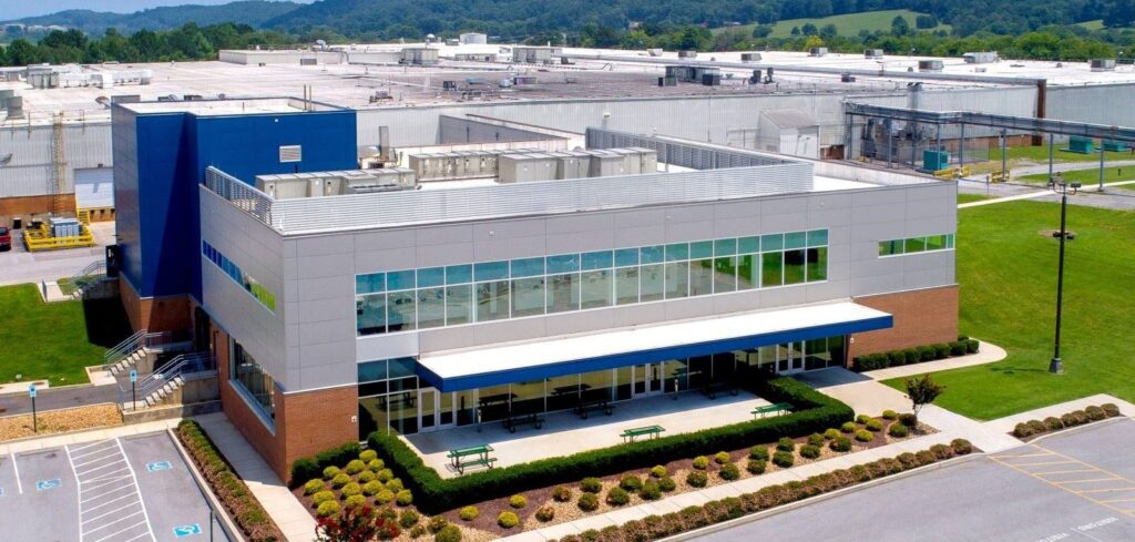 A modern industrial building with large windows, blue and white exterior, landscaped bushes, small trees, and benches out front; a large factory complex and green hills are visible in the background.