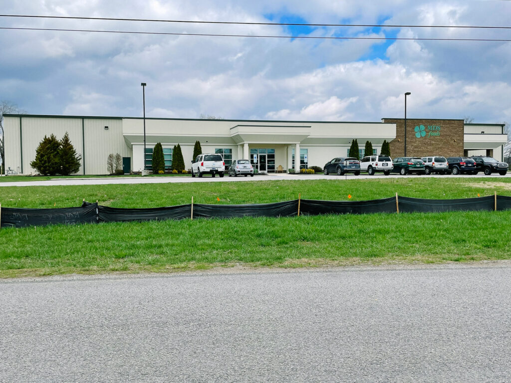 A single-story commercial building with a sign reading "MARS PETCARE." Several cars are parked in front, with green grass and a temporary black construction fence in the foreground under a partly cloudy sky.