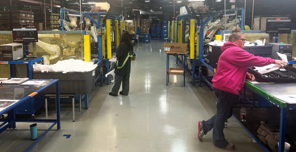 Two people work in a brightly lit factory or warehouse, standing at separate blue workstations with bins and papers. Yellow safety barriers and machinery are visible in the background.