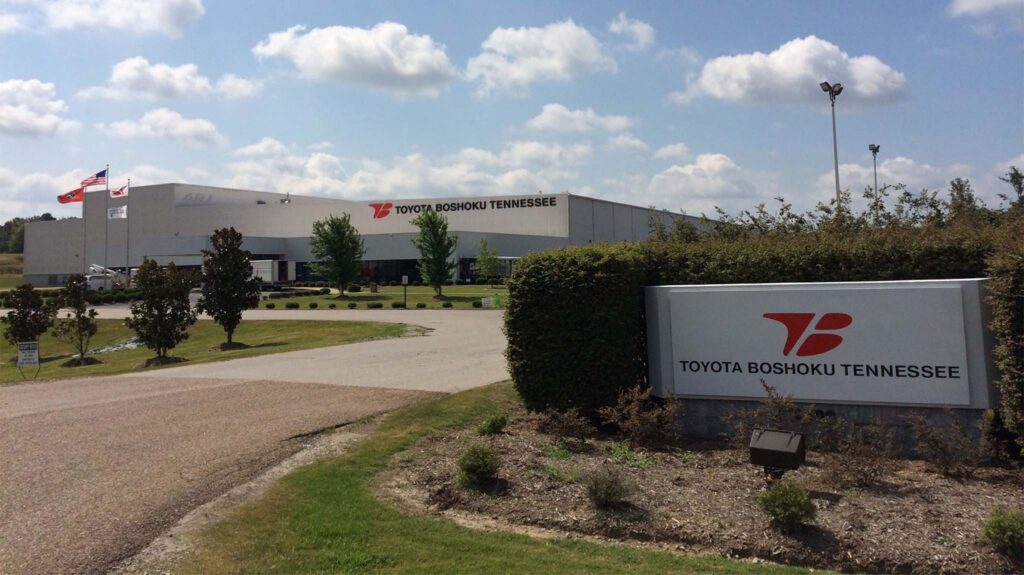A large industrial building with the sign "Toyota Boshoku Tennessee" at the entrance, surrounded by trees, grass, and an American flag flying on a pole. The sky is partly cloudy.