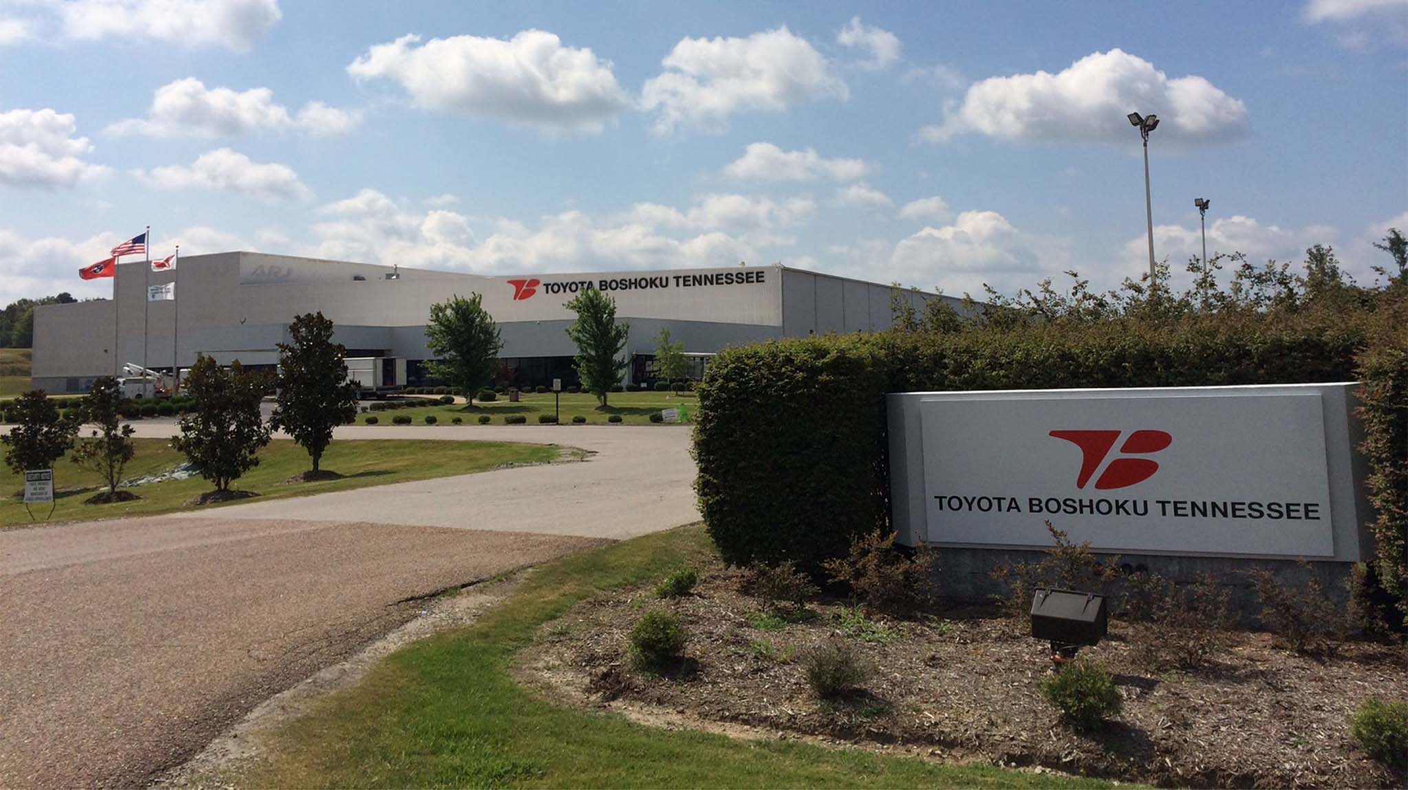 A large industrial building with the sign "Toyota Boshoku Tennessee" at the entrance, surrounded by trees, grass, and an American flag flying on a pole. The sky is partly cloudy.
