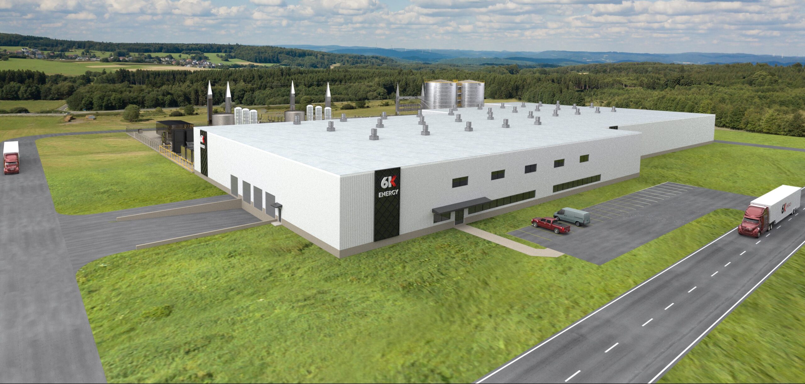 A large, modern white industrial building labeled "6K ENERGY" sits on green grass, with trucks parked nearby and a road leading to it. The landscape features trees, hills, and a partly cloudy sky in the background.