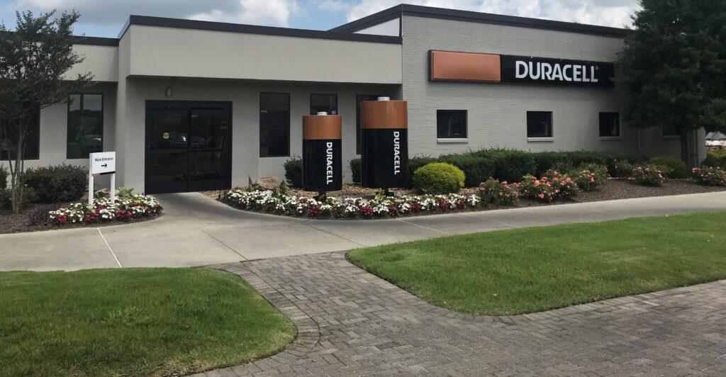A Duracell building with large battery-shaped displays outside, surrounded by landscaped bushes and flowers, with a sidewalk and a "Welcome" sign near the entrance.