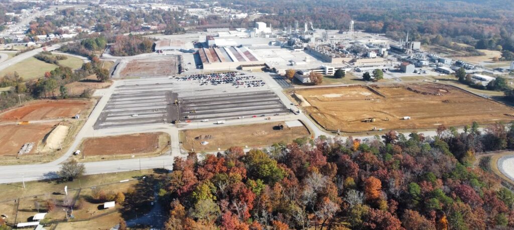 Aerial view of a large industrial facility with several parking lots, construction sites, and nearby wooded areas displaying autumn foliage. Roads border the site, and buildings are visible in the background.