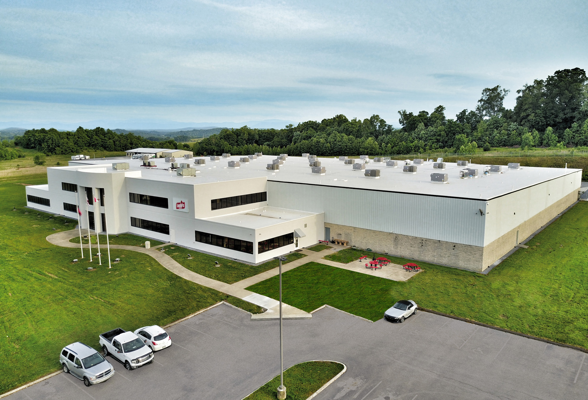 A large, modern white industrial or office building with flat roof, surrounded by green grass and trees. Several cars are parked outside, and red tables are set up near the entrance.