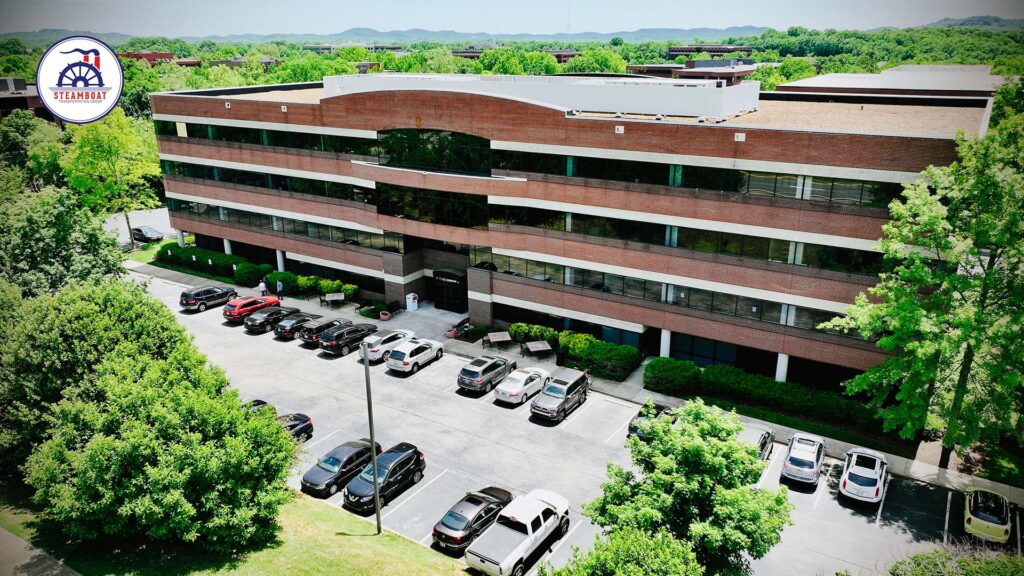 A large, four-story office building with a brick and glass exterior, surrounded by trees and a parking lot with several cars. The logo in the top left corner reads "STEAMAHAM.