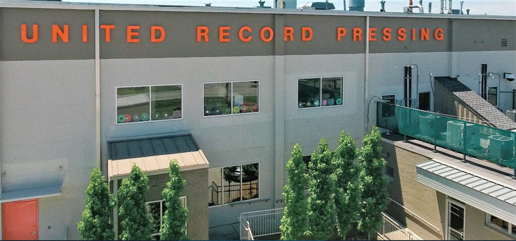 A large building with “United Record Pressing” in bold orange letters on the facade, featuring several windows, nearby trees, and a fenced outdoor area.