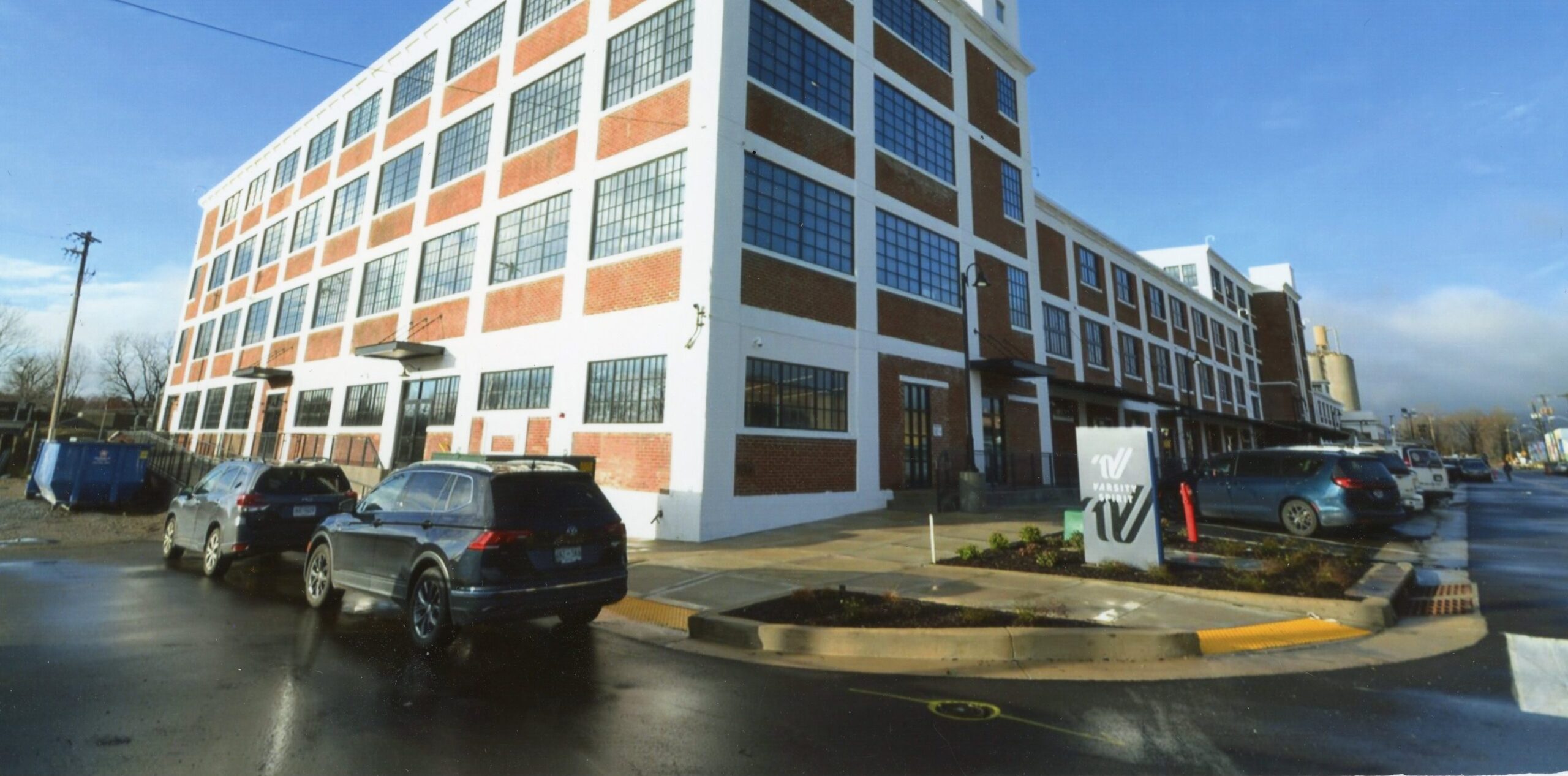 A large brick and white industrial-style building with many tall windows, cars parked along the wet street, and a sign in front displaying the letters "HV." The sky is partly cloudy.
