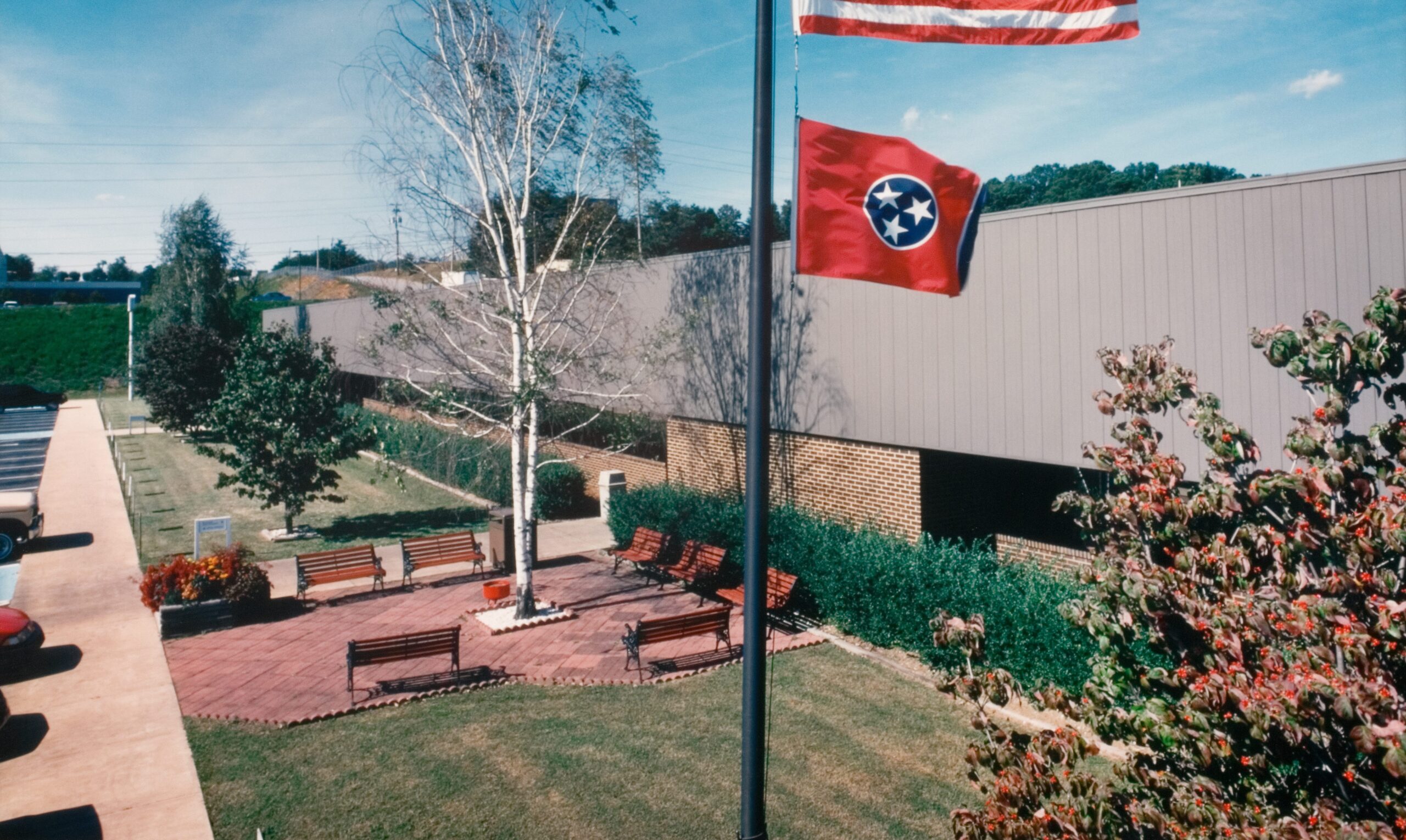 An outdoor courtyard with benches, trees, and a flagpole flying the Tennessee state flag and U.S. flag, next to a modern building under a blue sky.