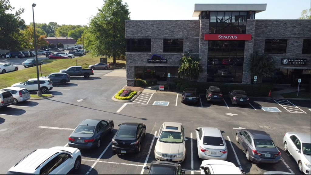 An office building with a Synovus sign, handicap parking spaces, and multiple cars parked in front. Trees and more parked cars are visible in the background on a sunny day.