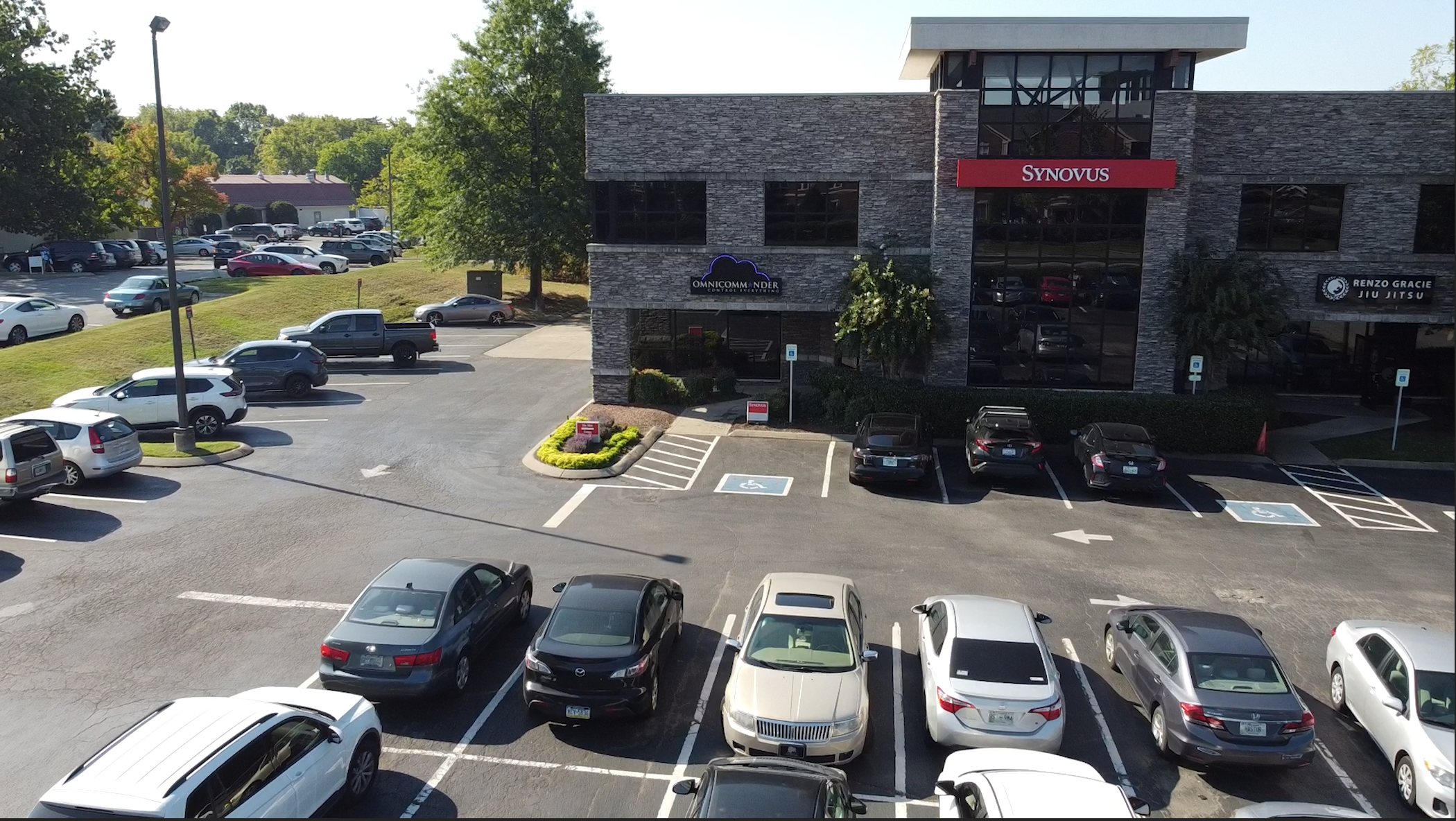 An office building with a Synovus sign, handicap parking spaces, and multiple cars parked in front. Trees and more parked cars are visible in the background on a sunny day.
