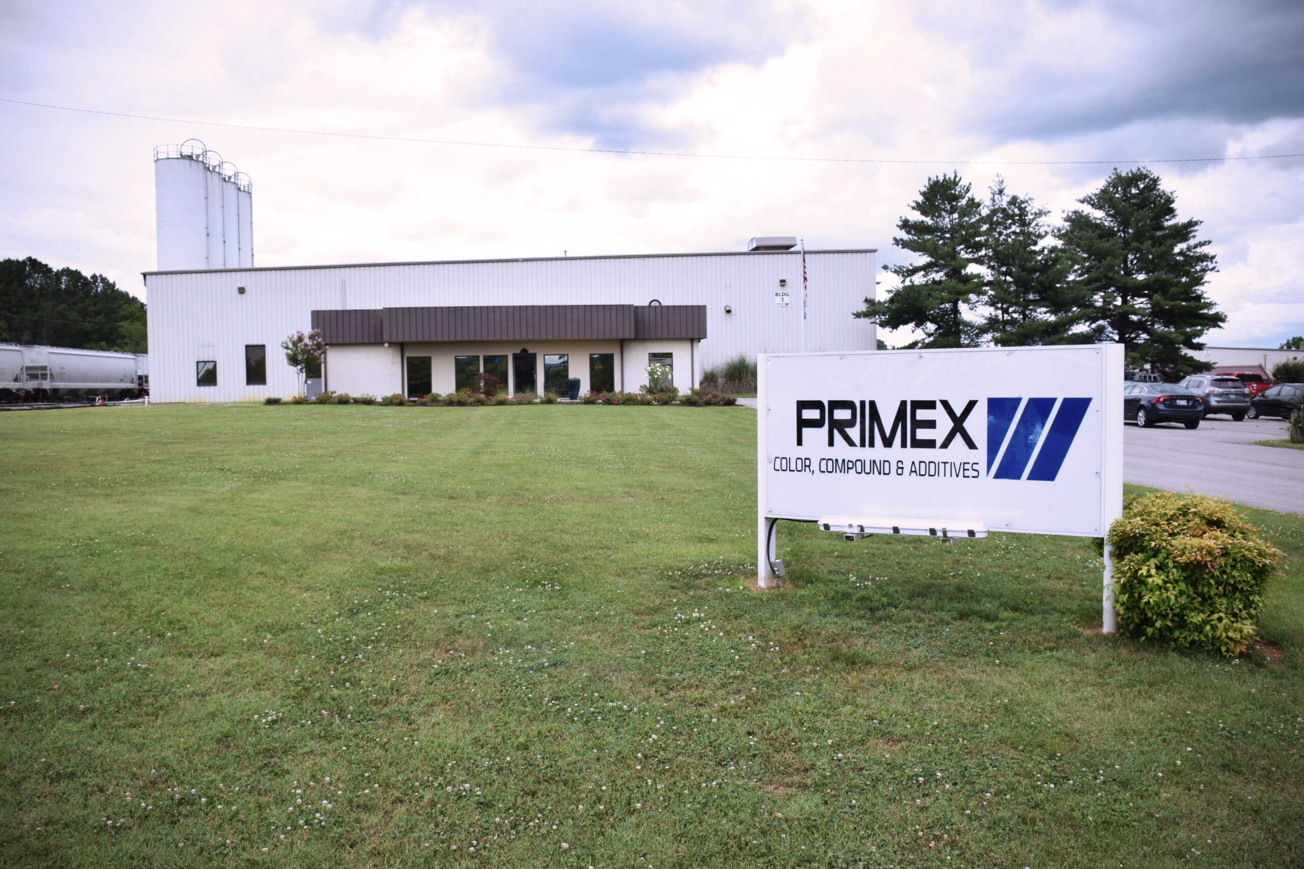 A large industrial building with a brown roof sits behind a well-kept lawn. In the foreground, a sign reads "PRIMEX Color, Compound & Additives." Several trees and parked vehicles are visible nearby.