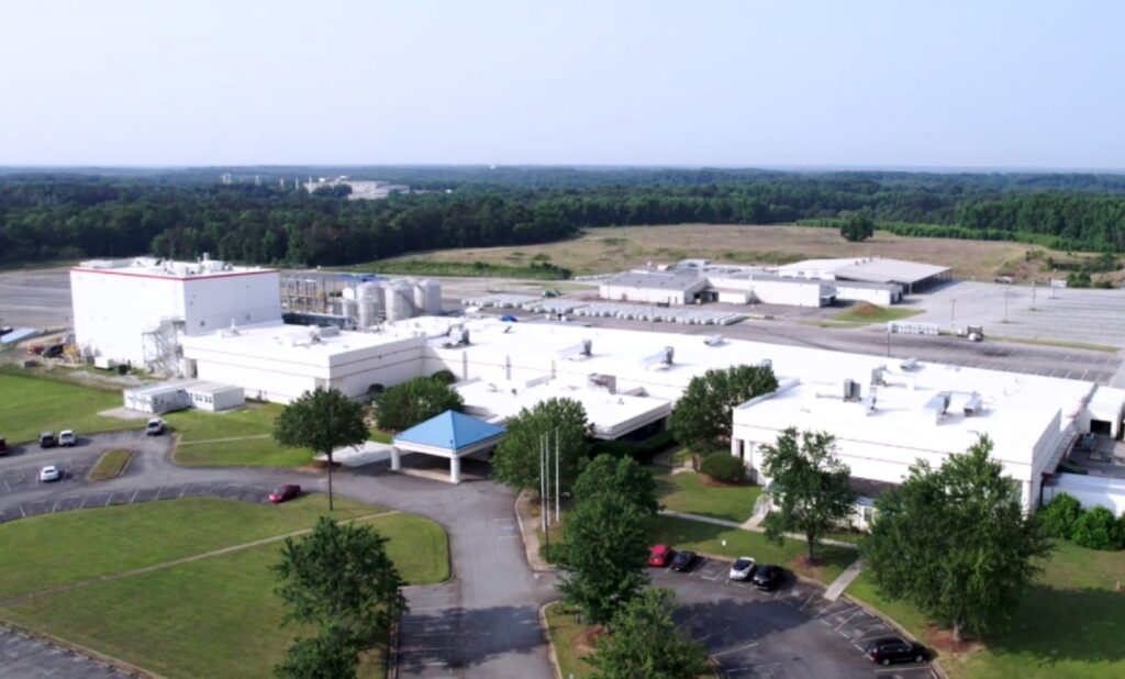 A large industrial facility with white buildings, surrounded by parking lots, grassy areas, and trees, set in a rural landscape with a forested background under a clear sky.