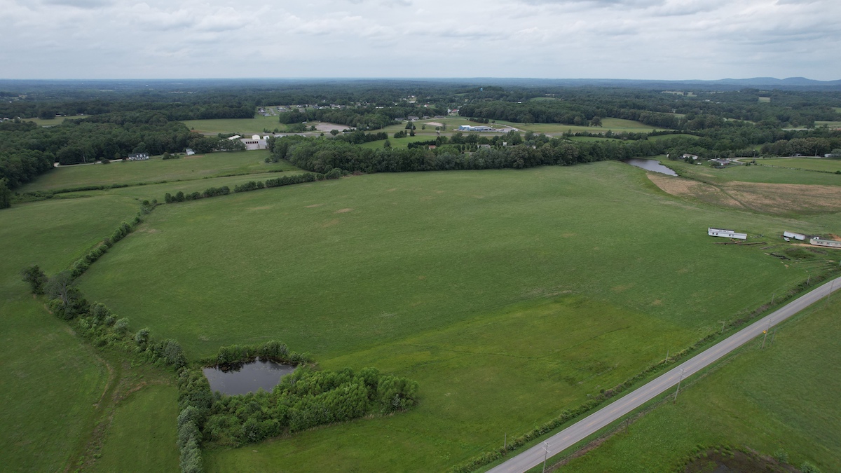 Aerial view of a large green field bordered by trees, a small pond in the foreground, and a road running along the right side, with scattered buildings and distant hills under a cloudy sky.