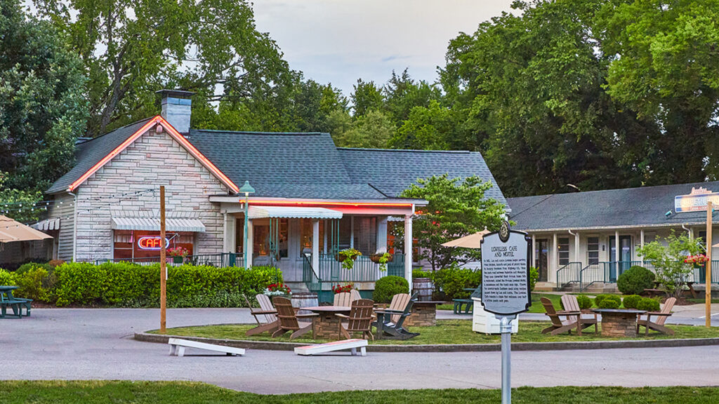 A small gray café with a neon sign sits beside a motel amid green trees. Wooden chairs circle fire pits on a grassy area, and cornhole boards are set up nearby. A historical marker stands in front of the café.