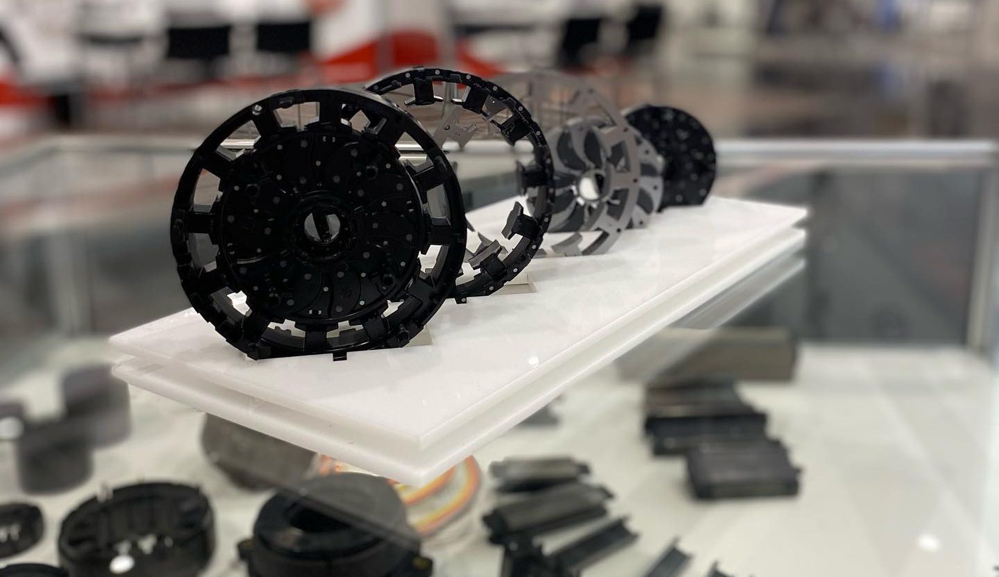 Various black and gray circular mechanical components are displayed on a white stand inside a glass case at an exhibition or trade show. Other industrial parts are visible in the background.