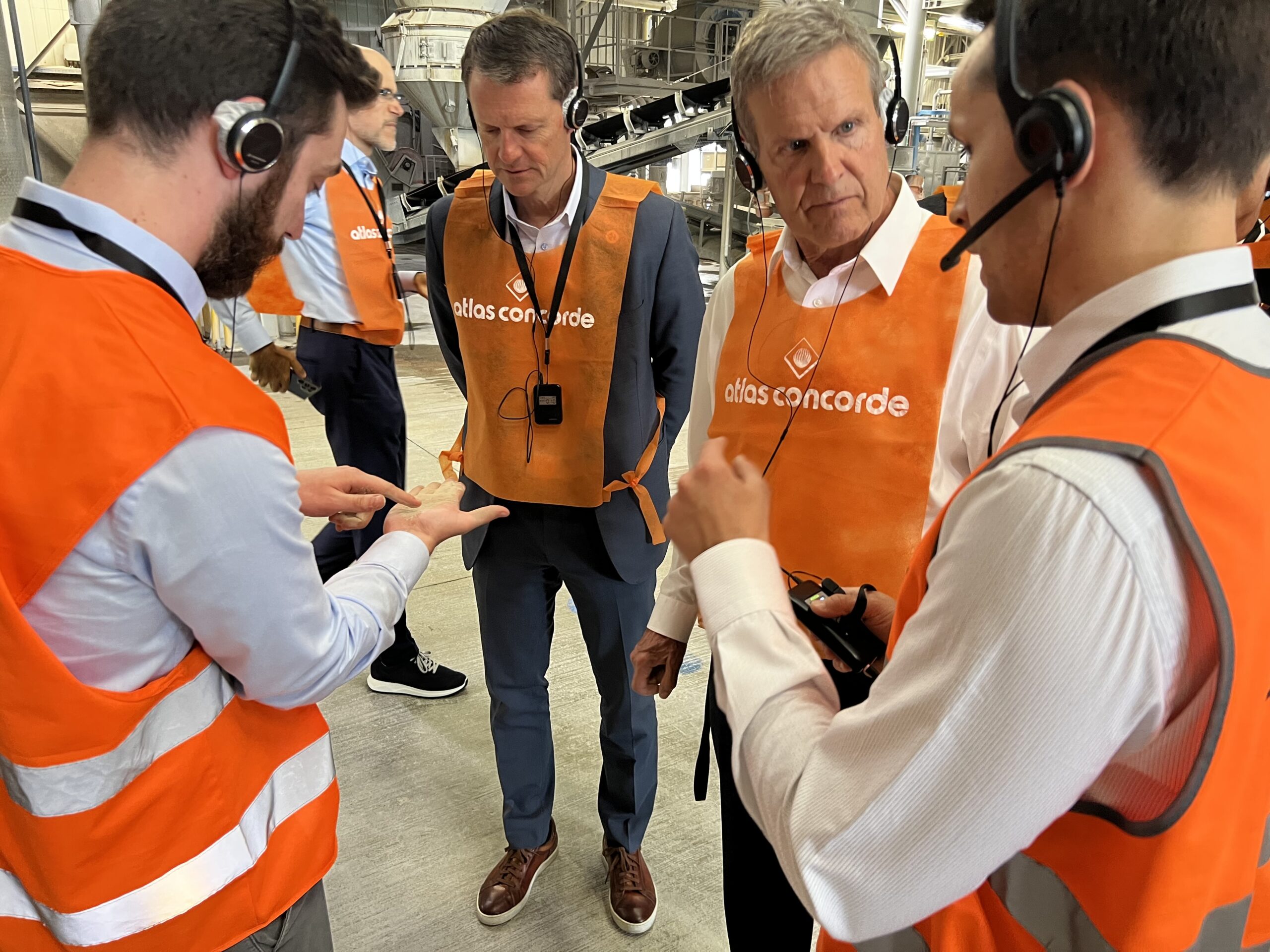 Four men wearing orange safety vests and headsets stand in a factory, with two of them intently discussing something as one points to his hand. Machinery and industrial equipment are visible in the background.