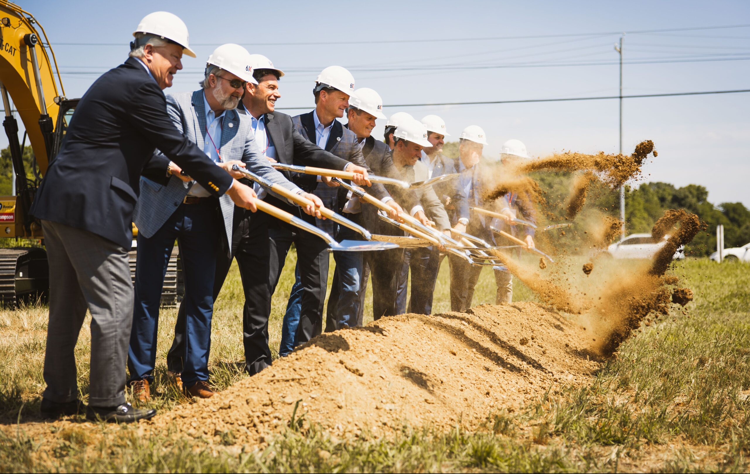 A group of people in suits and white hard hats use shovels to lift dirt at a groundbreaking ceremony on a sunny day, with an excavator and grass visible in the background.