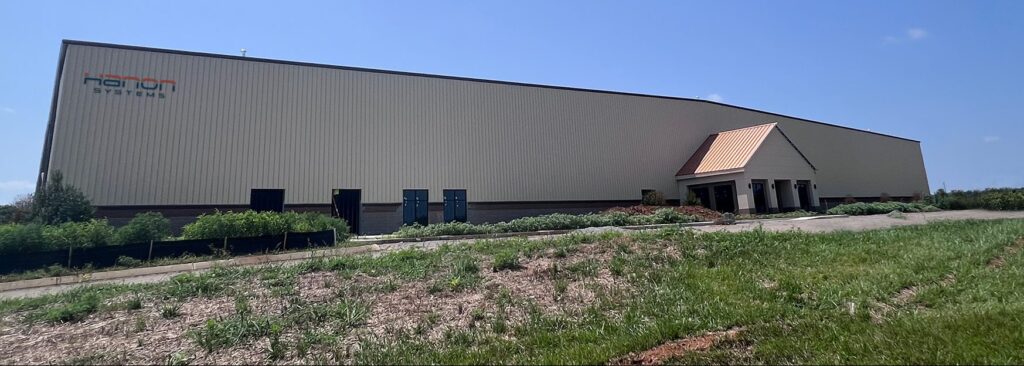 Large beige industrial building with a sloped roof, labeled "Hanon Systems" on the left side. The structure has a small entrance with columns and is surrounded by grass, shrubs, and a clear blue sky.
