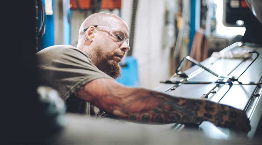 A man with glasses and tattooed arms works intently on a machine in an industrial workshop, surrounded by tools and equipment.