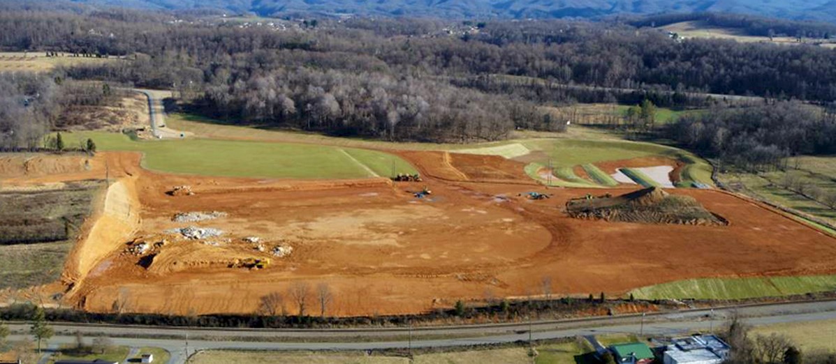 Aerial view of a large construction site with exposed reddish soil, construction vehicles, grassy patches, and a backdrop of leafless trees and rolling hills under a clear sky.