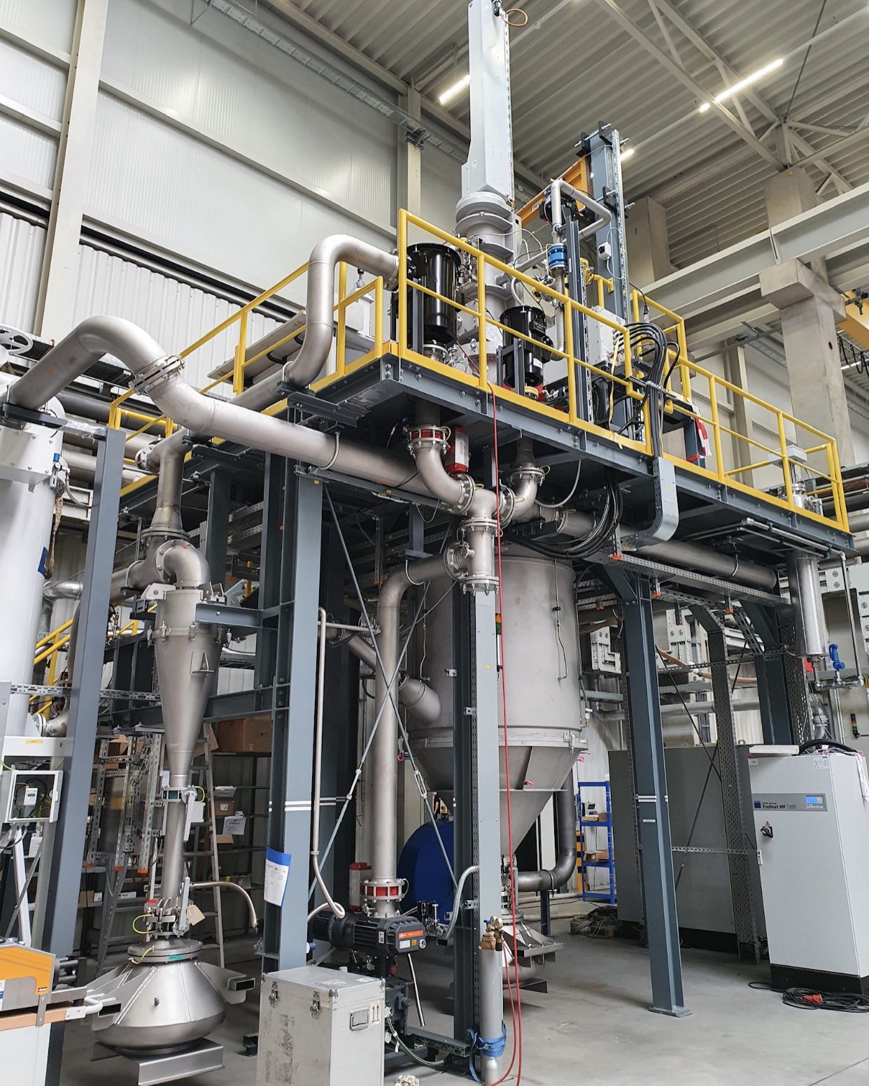Large industrial machine inside a factory, featuring metal pipes, tanks, and a platform with railings. The setting has high ceilings, bright lighting, and various equipment and control panels around the system.