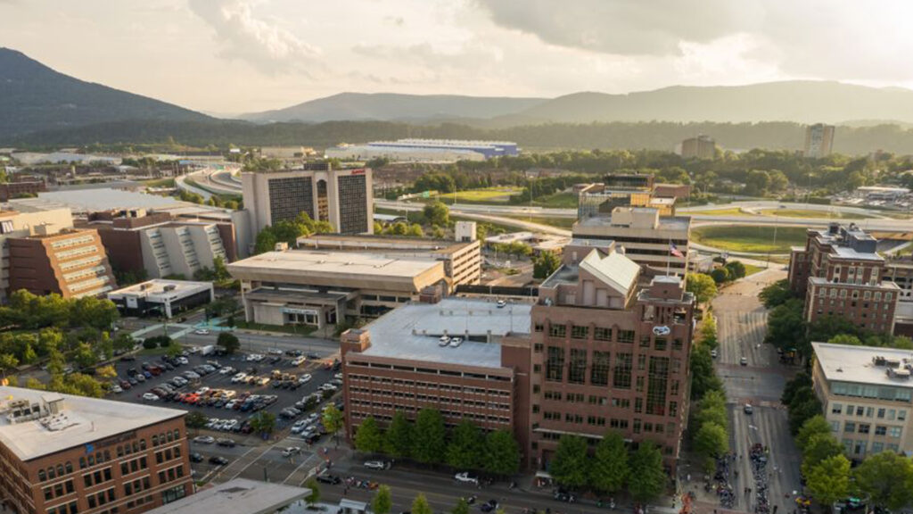 Aerial view of a city with office buildings, parking lots, tree-lined streets, and mountains in the background under a partly cloudy sky.