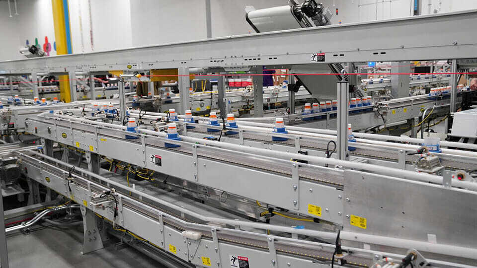 Rows of prescription pill bottles travel along automated conveyor belts inside a modern, brightly lit pharmacy or medication distribution facility.