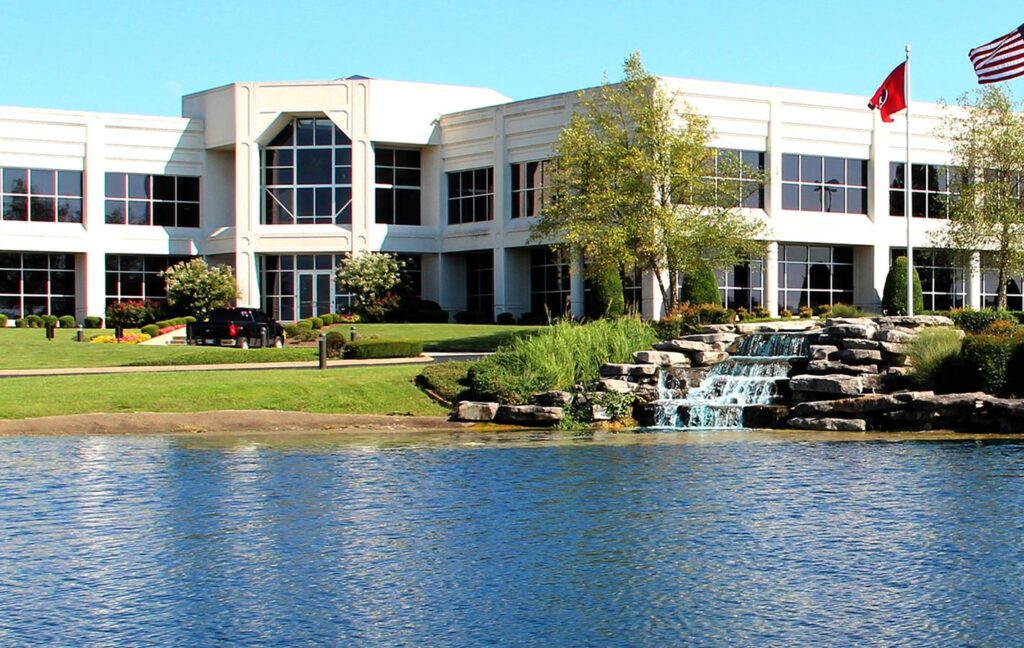 A modern office building with large windows sits behind a small pond and waterfall, surrounded by landscaped greenery and trees. Two flags are visible near the building under a clear blue sky.