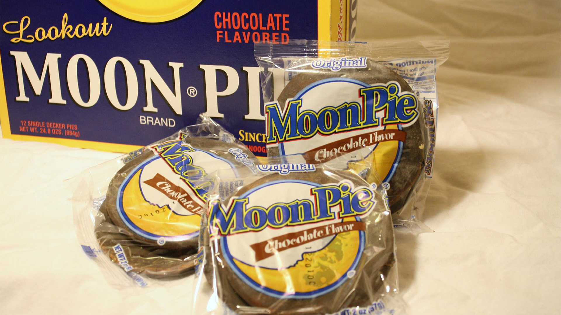 Three individually wrapped chocolate-flavored Moon Pie snacks are displayed in front of a large box labeled “Lookout Moon Pie Brand Chocolate Flavored Snacks.” The treats and packaging are arranged on a white surface.
