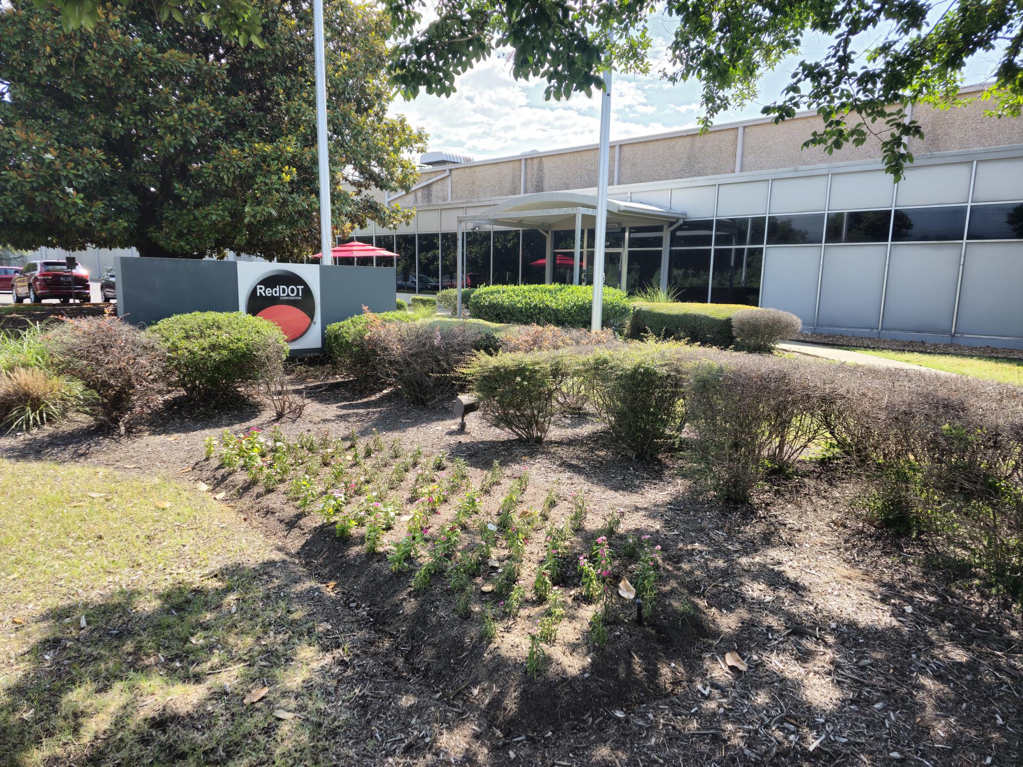 A landscaped area with bushes, small plants, and a sign reading "RedQDT" in front of a modern building with large windows and outdoor red umbrellas. Trees and a parked car are visible in the background.