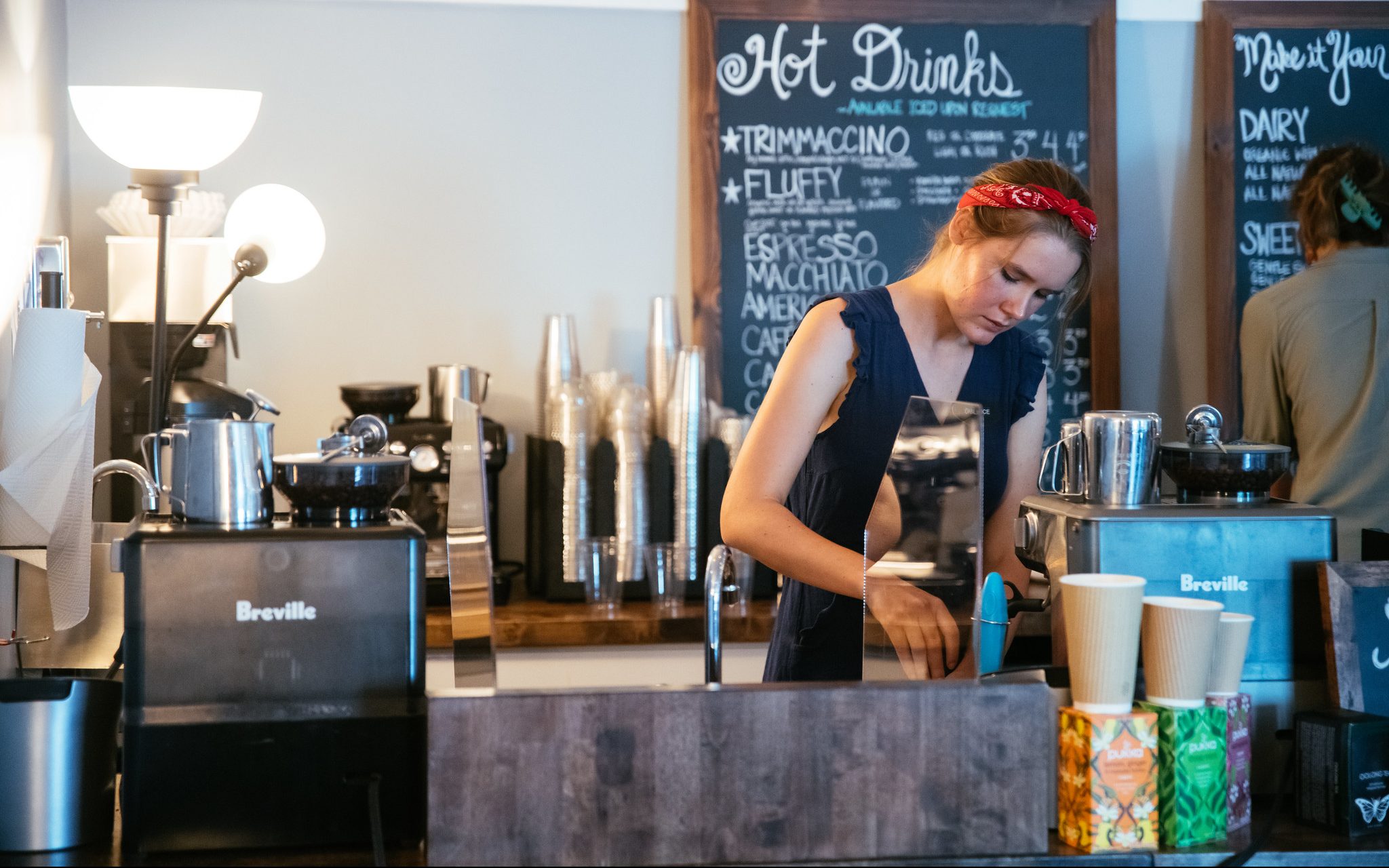 A barista in a red headband prepares a drink behind a coffee shop counter, with coffee machines, cups, and a chalkboard menu listing hot drinks in the background.
