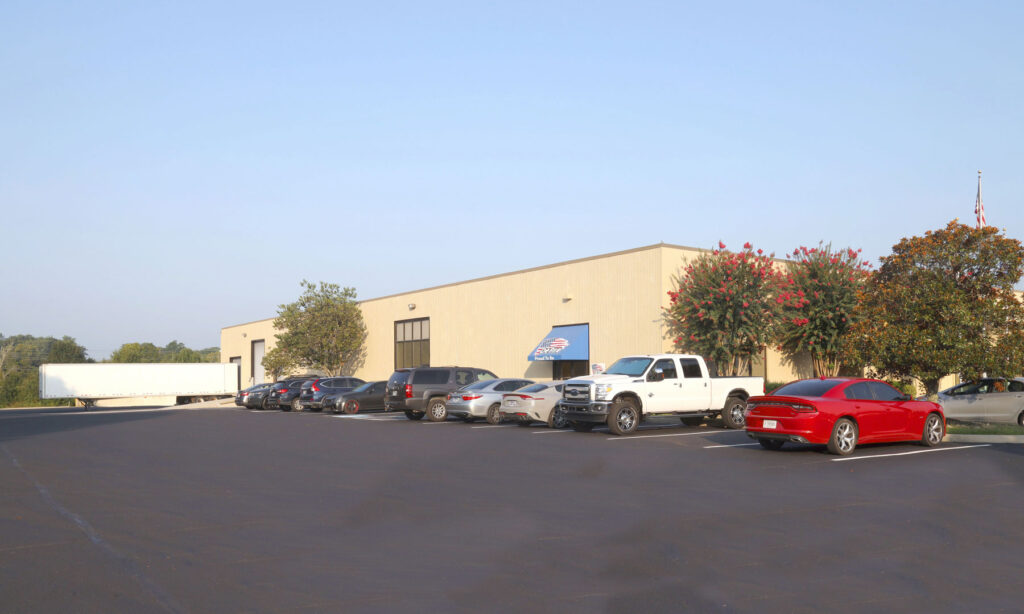 A row of cars and trucks is parked outside a large, beige industrial building with a loading dock and trees along the side, under a clear blue sky.