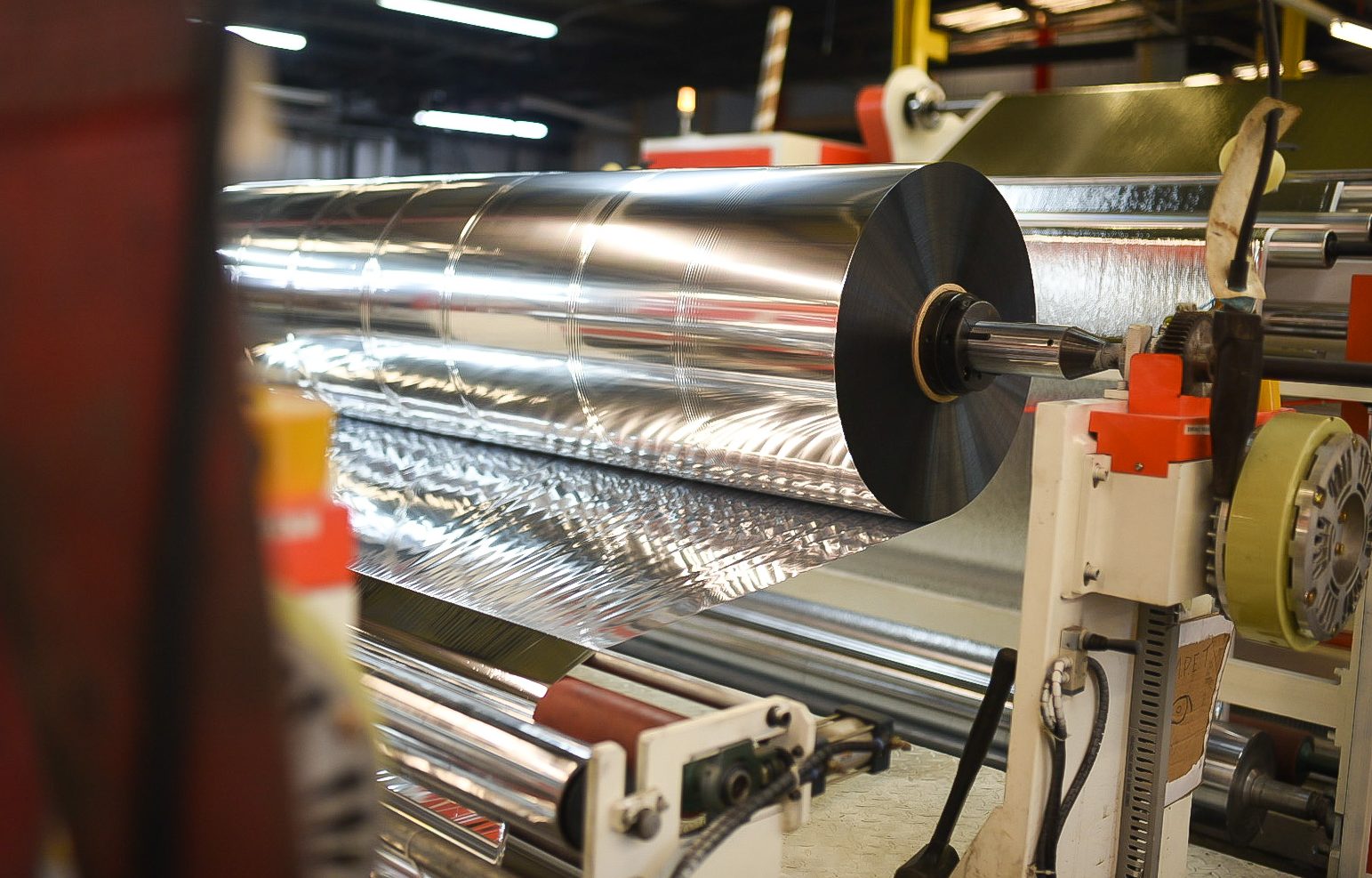 A large roll of shiny metallic foil being unwound and processed by industrial machinery inside a factory, with reflective surfaces and mechanical components visible.