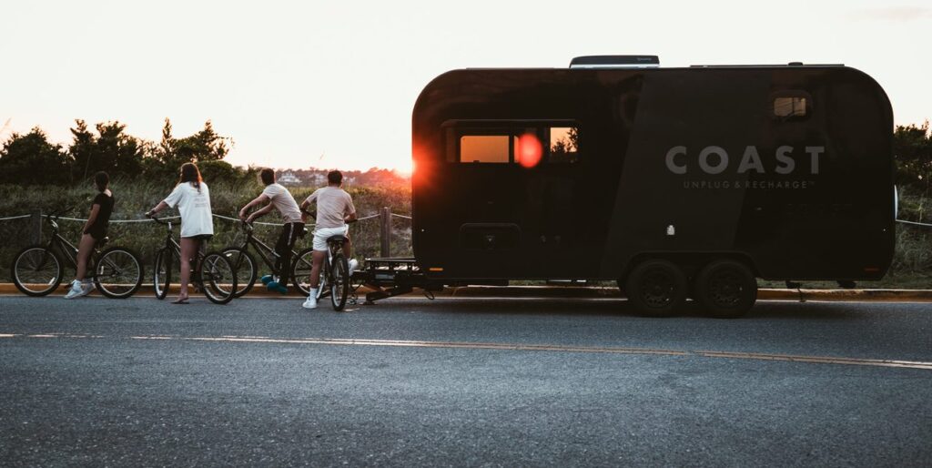 Four people with bicycles stand beside a black camper trailer at sunset. The trailer has "COAST" and "Unplug & Recharge" written on it. Trees and grass are in the background, and the road is in the foreground.