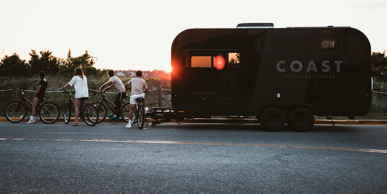 Four people with bicycles stand beside a black camper trailer at sunset. The trailer has "COAST" and "Unplug & Recharge" written on it. Trees and grass are in the background, and the road is in the foreground.