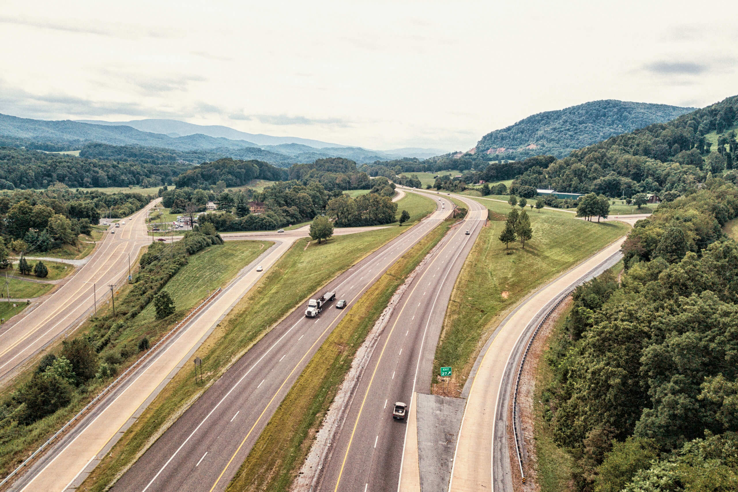 Aerial view of a highway with multiple lanes and vehicles driving, surrounded by green trees, grassy areas, and distant hills under a cloudy sky.