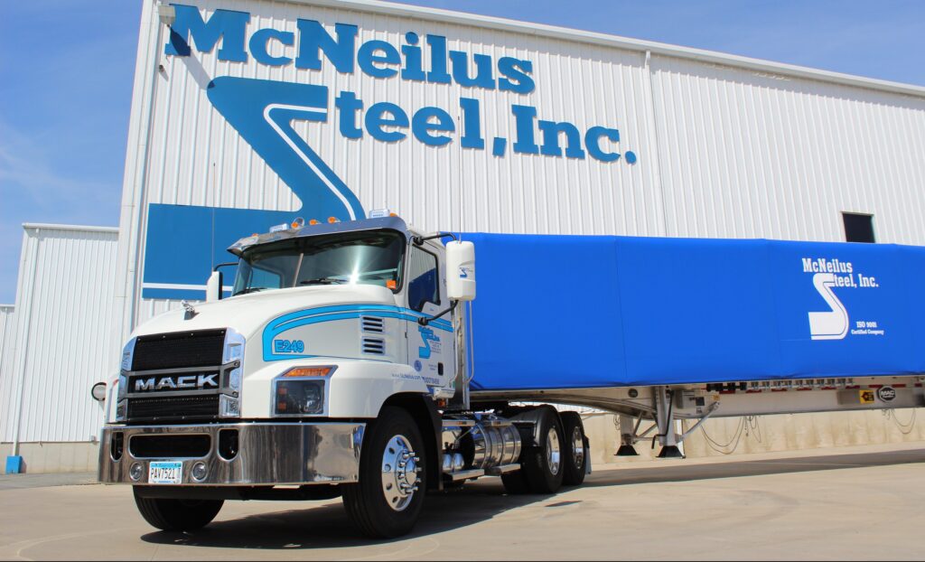A white and blue Mack semi-truck with a McNeilus Steel, Inc. trailer is parked in front of a large white building with the McNeilus Steel, Inc. logo on the side. The sky is clear and sunny.