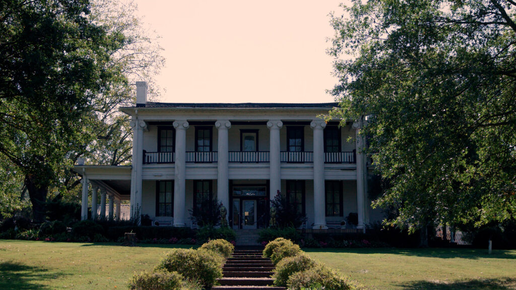 A large, two-story white mansion with tall columns, a covered porch, and a balcony, surrounded by trees and green lawns, with stone steps leading up to the entrance.
