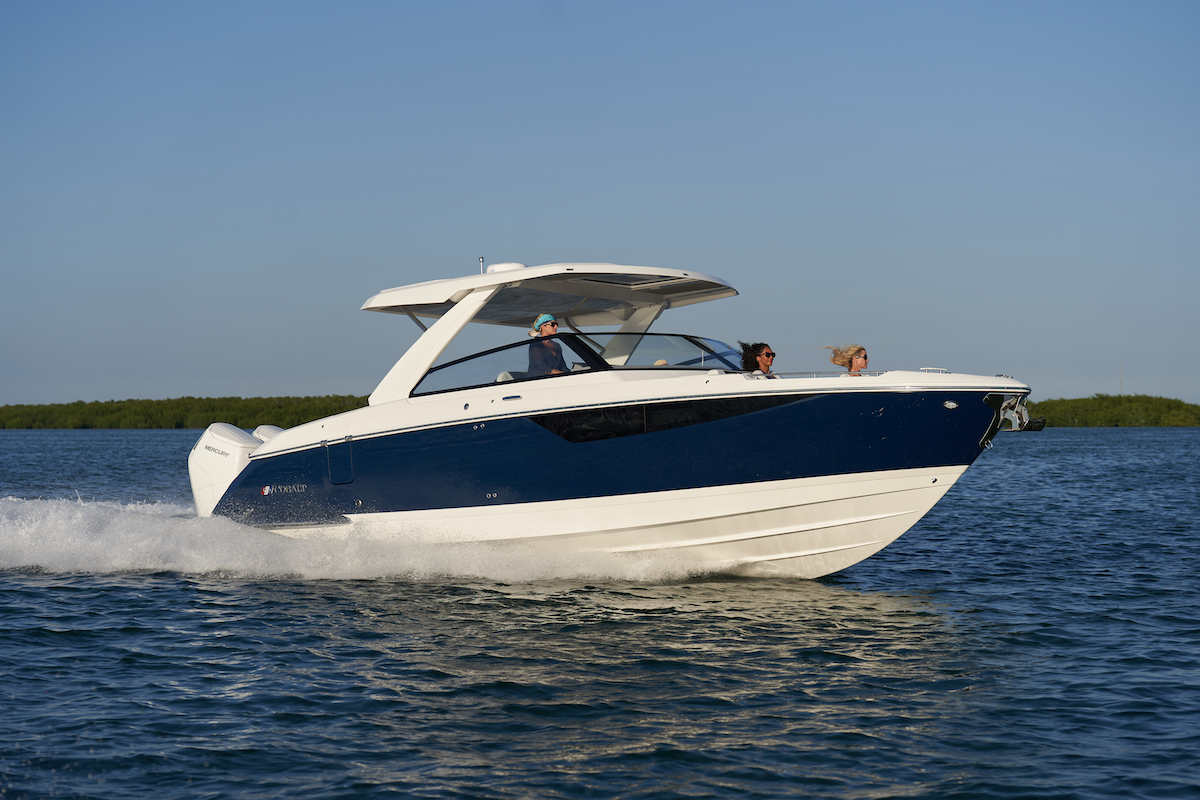 A modern blue and white motorboat speeds across calm water with three people on board under a clear blue sky, with green trees visible in the background.