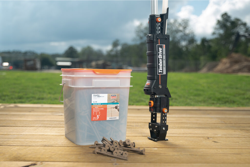 A black TimberDrive tool stands vertically on a wooden deck next to a plastic container filled with screws, some of which are scattered on the deck. The scene is outdoors with grass and trees in the background.