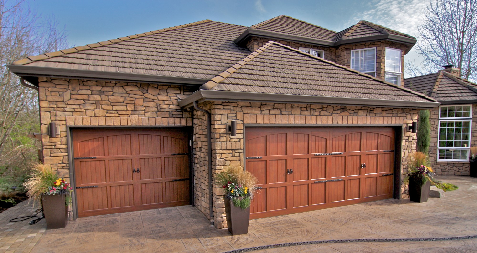 A stone-clad house with a two-car garage featuring wooden doors. Tall planters with flowers and ornamental grasses stand on each side of the garage. The driveway is stamped concrete, and the roof is covered with brown shingles.